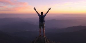 Person on mountain peak at sunrise, arms raised.